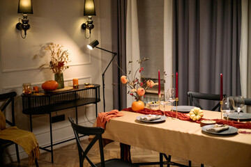 Empty dining room table set for Thanksgiving dinner with plates, wine glasses, candles, autumn decorations, pumpkins and floral centerpiece, chairs surrounding table, cozy indoor setting