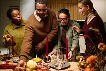 Diverse group of young adult and middle aged men and women gathering around table serving traditional Thanksgiving dinner, smiling and interacting while reaching for food together