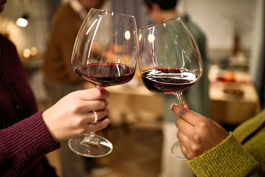 Unrecognizable female friends clinking wine glasses during Thanksgiving dinner, toasting each other, hands visible in foreground, people socializing in background