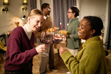 Young adult Caucasian woman and young adult Black woman clinking wine glasses and smiling, multiethnic group of young adults in background serving food during Thanksgiving dinner among friends