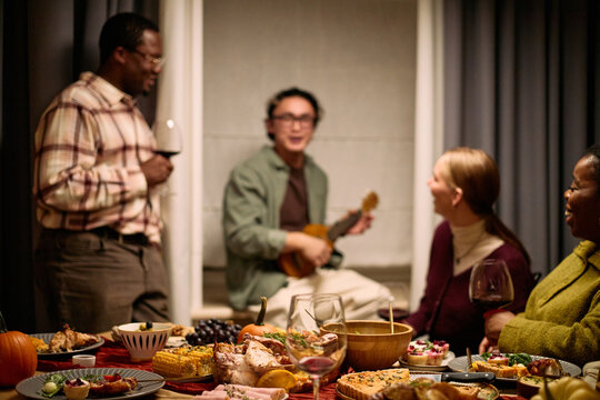 Group of diverse young adults and middle aged people gathering around table sharing Thanksgiving dinner, Black man standing holding wine glass, Asian man playing guitar, Caucasian woman smiling