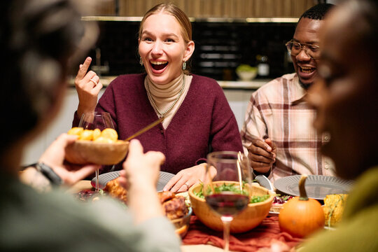 Diverse group of young adults and middle aged people laughing and sharing food around table during Thanksgiving dinner, Caucasian woman smiling, Black man enjoying meal, hands passing dishes