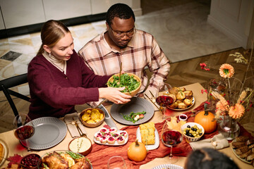 Young Caucasian woman serving salad to young Black man during thanksgiving dinner among friends, multiethnic couple sitting at table sharing traditional holiday food and drinks