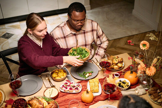 Young Caucasian woman serving salad to young Black man during thanksgiving dinner among friends, multiethnic couple sitting at table sharing traditional holiday food and drinks