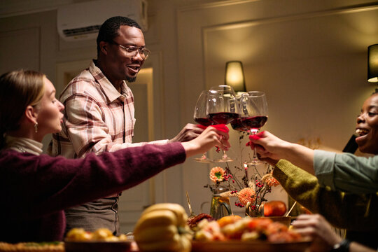 Group of diverse young adults and middle aged Black man celebrating Thanksgiving dinner among friends, raising wine glasses together at table with festive food and autumn decorations