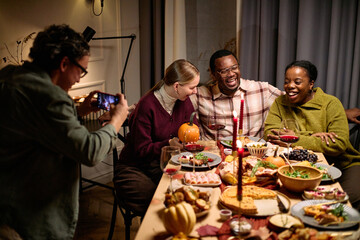 Group of diverse young adults and middle aged friends sitting around table sharing Thanksgiving dinner, smiling and laughing while man photographing moment with smartphone