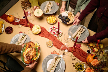 Diverse group of young adult and middle aged men and women serving traditional Thanksgiving dinner, arranging turkey, fruit, snacks and desserts on festive table, hands visible from above