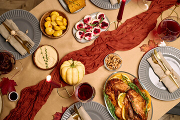 Overhead view showing festive Thanksgiving dinner table with roasted turkey, assorted side dishes, desserts, wine glasses, candles, autumn leaves, plates, cutlery, napkins