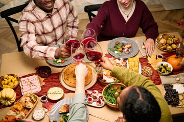 Diverse group of young adult and middle aged men and women friends clinking wine glasses over Thanksgiving dinner table, sharing meal with traditional dishes, celebrating friendship together