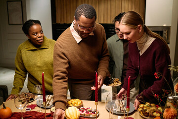Obraz premium Group of young adult multiethnic friends gathering around table serving traditional Thanksgiving dinner food, Black man reaching for dish while Caucasian woman and others smiling and interacting
