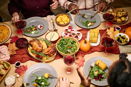 Diverse group of young adult and middle aged men and women sharing Thanksgiving dinner, passing plates and serving food at festive table with roasted turkey, vegetables, salads, wine glasses