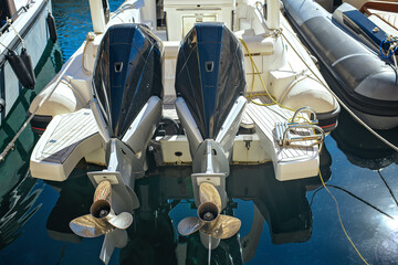 Close-up of two outboard marine engines on a modern motorboat in a marina, with deck details, propellers, and water reflections on a bright sunny day.