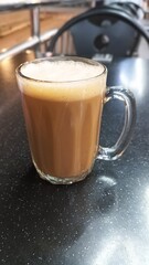 A glass mug of frothy teh tarik served on a dark tabletop.