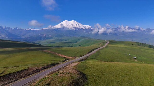 The car is driving along a mountain road in the Caucasus, towards the foot of Mount Elbrus in the Dzhily-Su tract, with the snow-capped peak of Mount Elbrus on the horizon