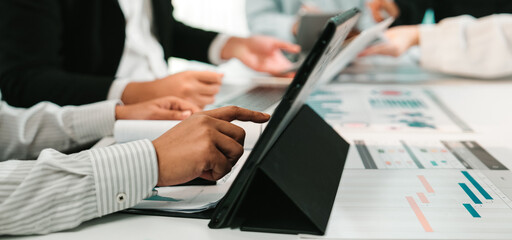 A diverse group of business professionals engaged in a meeting, analyzing data displayed on a tablet, surrounded by charts and documents, emphasizing collaboration and productivity. SACTR
