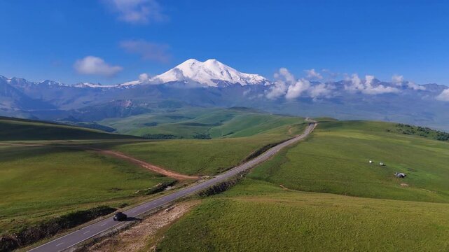 The car is driving along a mountain road in the Caucasus, towards the foot of Mount Elbrus in the Dzhily-Su tract, with the snow-capped peak of Mount Elbrus on the horizon