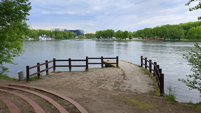 On the shore of the lake there is a tiled pier with a wooden fence, alder and birch trees grow. On the far shore there is a park with trees and buildings behind it. There are ripples on the water