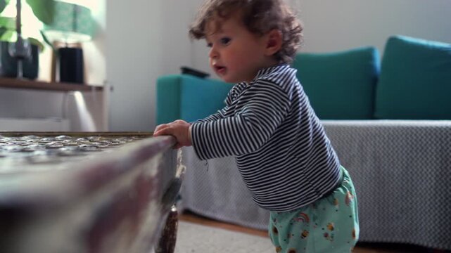 Curious toddler steadying body while rising to stand beside wooden table in cozy living room, conveying early balance, determination, and exploration of home environments