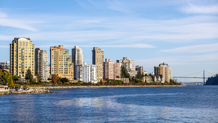 Obraz premium A bright waterfront skyline featuring tall residential towers along a rocky shoreline. Calm blue water, clear sky, and modern urban architecture create a vibrant coastal city scene ideal for travel