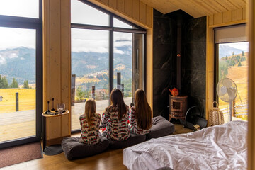 Family in cozy cabin enjoying Mountain View. Three girls in matching pajamas sit by a large window in a wooden cabin, peacefully enjoying a scenic mountain landscape.