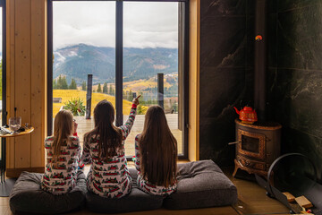 Children in pajamas pointing at Mountain View. Three girls in matching festive pajamas sit on cushions in a cozy cabin, pointing out at a scenic misty mountain landscape.