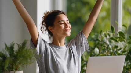 Young woman with brown hair, wearing a gray t-shirt, is celebrating success with arms raised in a bright indoor space filled with greenery and a laptop on the table, conveying joy and accomplishment