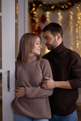 Young Caucasian couple, man and woman in knitted sweaters and jeans, embracing and smiling indoors, warm fairy lights and festive decor in background. Concepts: love, winter, romance, holiday. 
