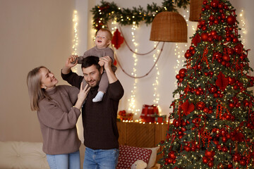 Young Caucasian parents with baby, smiling, father holding child on shoulders in cozy living room with Christmas decor and white wall. Concepts: family, celebration, warmth, holidays.