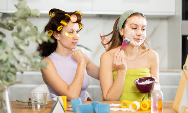 Carefree relaxed adult woman wearing headband, wrapped in towel, enjoying home spa day with bestie, applying facial mask with brush during joint beauty ritual