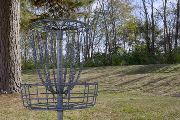 A metal Disc Golf basket (hole) with chains, set on a grassy course with trees and a pine trunk nearby on a sunny day.