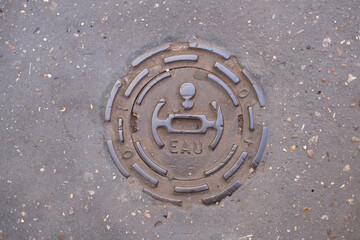 Round metal utility cover labeled “EAU” (“Water”) set in textured asphalt.