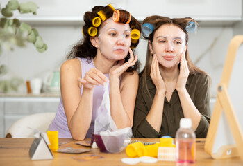Two female friends sharing moment of relaxation and enjoying spa day at home, sitting by tabletop vanity mirror with colorful rollers in hair and applying moisturizing sheet masks to faces