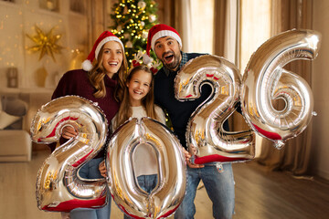 Overjoyed family celebrating Christmas at home, having fun holding giant balloons shaped as numbers 2026