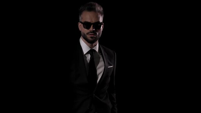 Serious man in sunglasses and suit is looking down, then looking away, and finally buttoning his jacket on dark studio background.