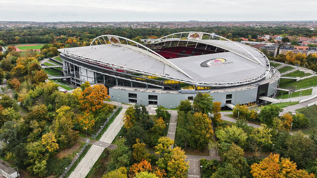 Telephoto view of RB Leipzig Arena centered, framed by trees with colorful autumn leaves, other city districts visible in the background.