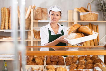 Female baker worker sells loafs of breads. Authors bakery, fresh pastry. In sales hall near showcase, she demonstrates finished products, lures customers with fragrant loafs of bread