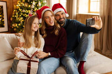 Happy European family dressed in festive clothes taking selfie or video calling, sitting on sofa in room with Christmas tree. Winter holiday season