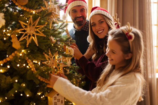 Parents and daughter having fun decorating Christmas tree, placing Christmas lights and ornaments on it, decorating home for winter holiday season - Powered by Adobe