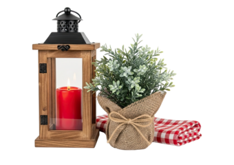 A rustic wooden lantern with a burning red candle, a burlap-wrapped plant, and a checkered napkin create a cozy and warm decorative still life composition isolated on a black background