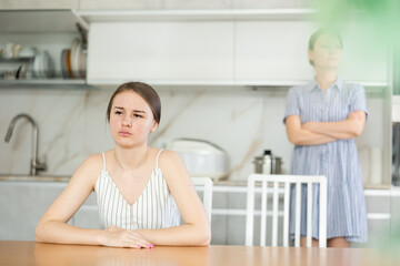 Upset teenage girl sitting at the table turning her back upon mother standing crossly at the...