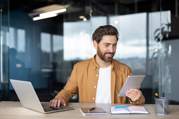 Businessman using a laptop and a digital tablet, sitting at a desk in a contemporary office, focusing on his work with a positive and engaged expression