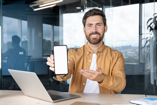 Businessman sitting at office desk smiling and holding up a smartphone with a blank white screen for app or website mockup, laptop and workspace in background