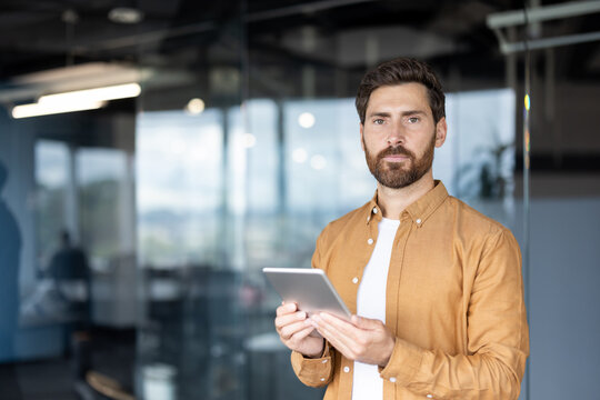 Young businessman holding a digital tablet with a serious expression, looking directly at the viewer, standing in a contemporary office representing digital communication and business technology - Powered by Adobe
