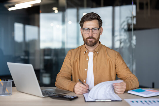 Businessman looking at camera, sitting at office desk, holding documents, writing on clipboard, with laptop and calculator in a modern office environment