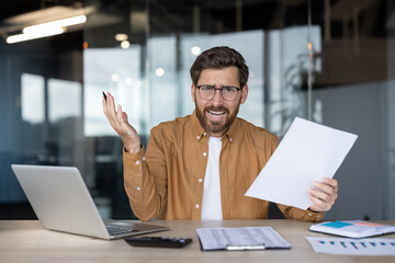 Young man in glasses sitting at office desk feeling frustrated about business problems, holding up a document and throwing his other hand in the air in annoyance