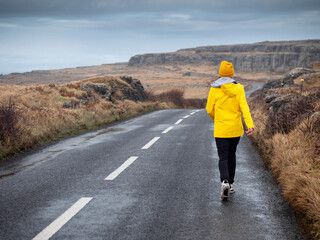 Fototapeta premium Teenager girl in yellow jacket walking on a small country road, picturesque nature scenery with rough stone terrain in the background. Burren, Ireland. Travel and tourism. Exploring Irish nature.