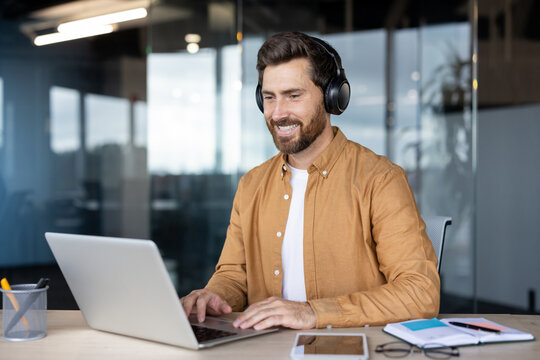 Happy bearded man in smart-casual attire wearing headphones, smiling at laptop during a virtual meeting or webinar in a bright modern office, focused and productive