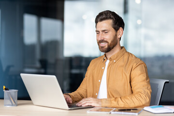 Smiling man concentrating and typing on a laptop, working productively from a modern office while looking at the screen, representing business success and remote work efficiency