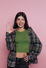 portrait of young lively girl showcasing a fun 'OK' gesture with a wink, radiating energy and positivity in her casual outfit on pink background