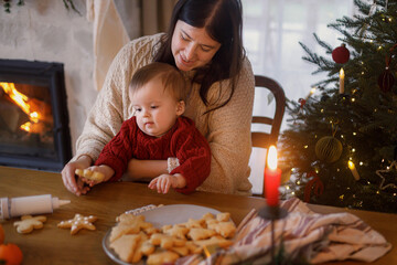 Happy mother with cute baby son making gingerbread christmas cookies together, decorating with sugar glaze against christmas tree. Christmas family traditions, winter holiday preparation